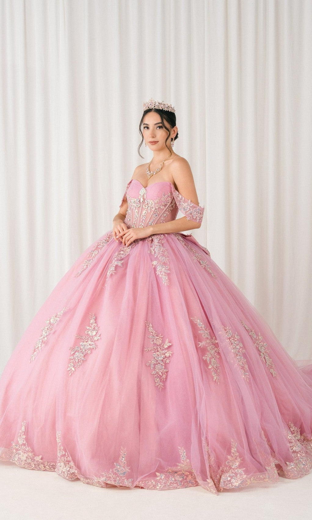 A young woman stands before a white curtain wearing the Quinceanera Dress 8405JQ by Cinderella Couture in Dusty Rose, an off-the-shoulder ball gown with delicate floral embroidery and a matching tiara.